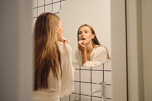 Jeune femme magnifique en robe blanche regardant rêveusement dans un miroir appliquant du rouge à lèvres dans la salle de bain seule