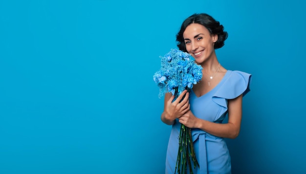 Une jeune femme magnifique vêtue d'une robe bleu vif regarde la caméra avec un grand sourire, tenant un bouquet de fleurs bleues dans ses mains. Journée internationale de la femme