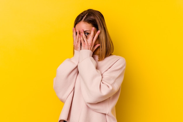 Jeune femme maigre caucasienne isolée sur un mur jaune clignote à travers les doigts effrayés et nerveux.