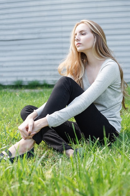 Jeune femme maigre de race blanche aux cheveux bruns assis sur une pelouse verte au mur