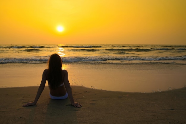 Jeune femme en maillot de bain blanc assis sur la plage au coucher du soleil