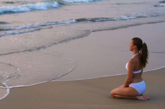 Jeune femme en maillot de bain blanc assis sur la plage au coucher du soleil