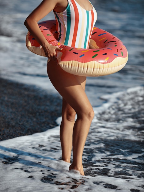 Jeune femme en maillot de bain debout sur une plage avec un anneau de beignet gonflable et profitant d'un bain de soleil.