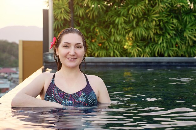 Jeune femme en maillot de bain avec fleur dans les cheveux mouillés sourit et regarde la caméra dans la piscine extérieure au coucher du soleil
