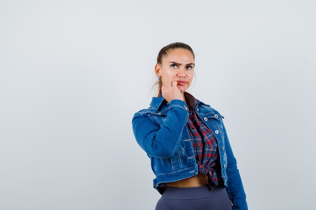 Jeune femme avec la main près de la bouche en chemise à carreaux, veste en jean et à la perplexité.