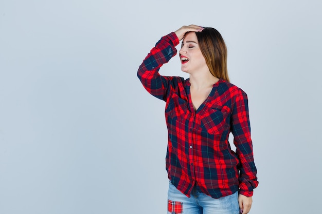 Jeune femme avec la main sur la tête, gardant les yeux fermés en chemise à carreaux, jeans et l'air harcelé, vue de face.