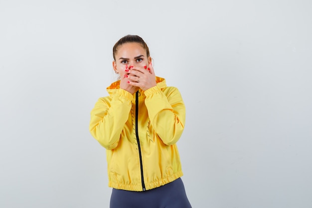 Jeune femme avec les mains sur la bouche en veste jaune et à la perplexité, vue de face.