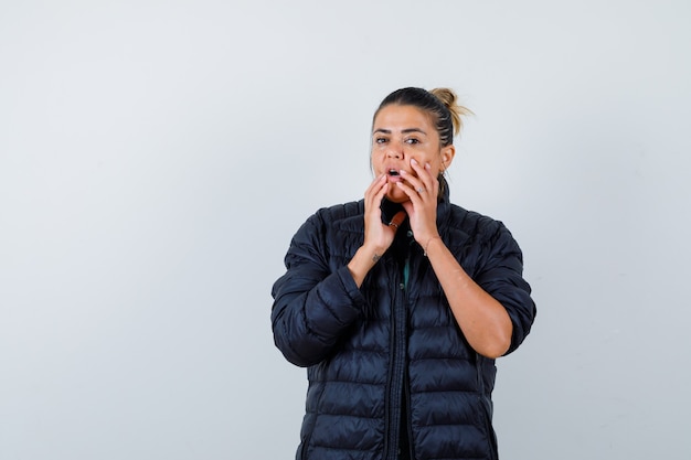 Jeune femme avec les mains près de la bouche ouverte en doudoune et l'air surpris, vue de face.