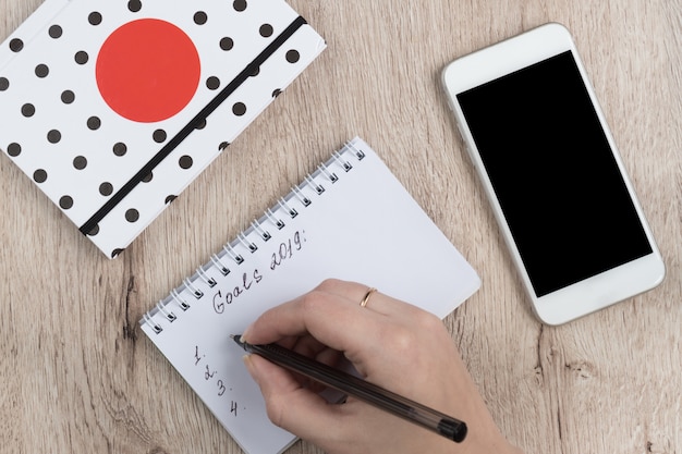 Jeune femme mains tenir pages ouvertes du cahier avec un stylo noir sur une table en bois. Liste des objectifs 2019.