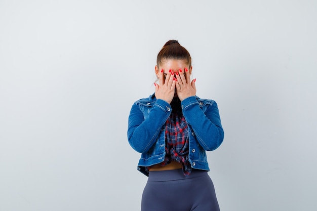 Jeune femme avec les mains sur le visage en chemise à carreaux, veste, pantalon et à la nostalgie. vue de face.