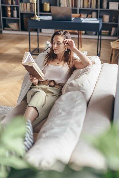 Jeune femme à la maison allongée sur un canapé et un livre de lecture.