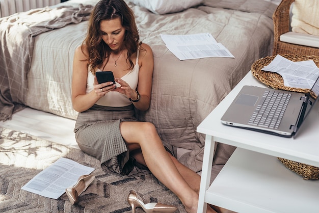 Jeune femme à la maison après le travail, a enlevé ses chaussures, est assise détendue sur le sol et utilise le smartphone.