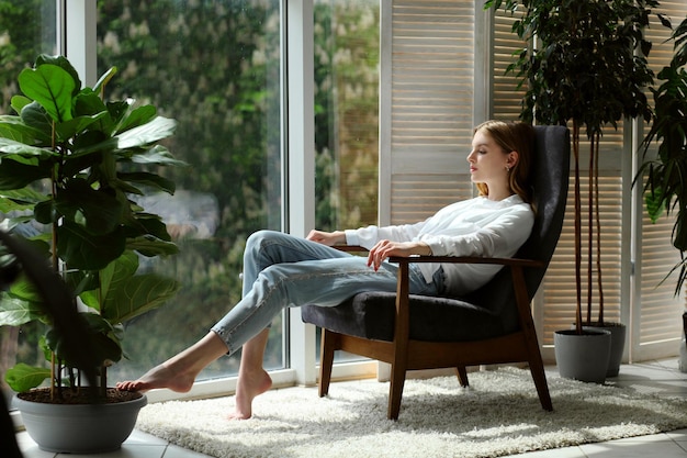 Jeune femme à la maison assise sur une chaise moderne près de la fenêtre se relaxant dans le salon