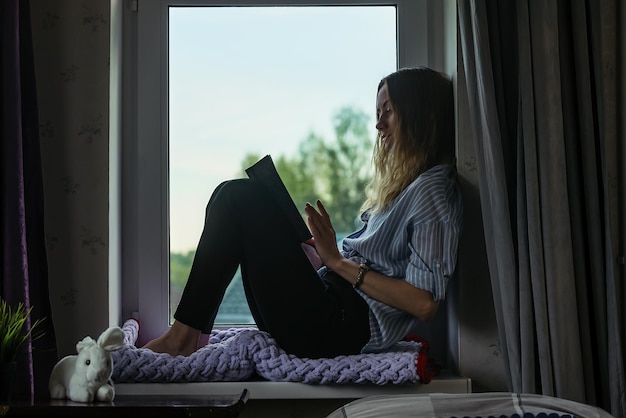 Jeune femme à la maison assise sur le rebord de la fenêtre et lisant un livre