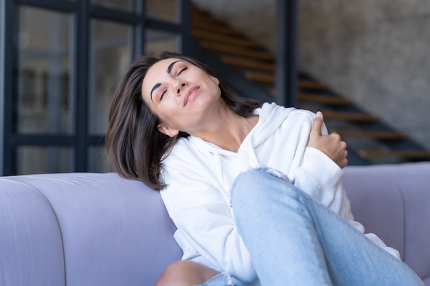 Une jeune femme à la maison dans un sweat à capuche blanc sur le canapé est assise paisiblement, aime la solitude