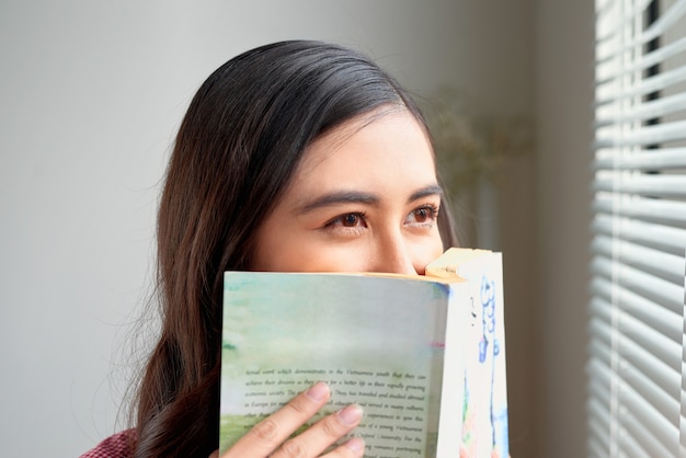 Jeune femme à la maison debout près de la fenêtre se relaxant dans son livre de lecture de salon.