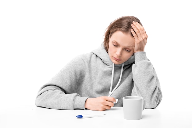 Jeune femme malade bouleversée en capuche assis par bureau, mesurer la température et prendre des médicaments tout en restant à la maison