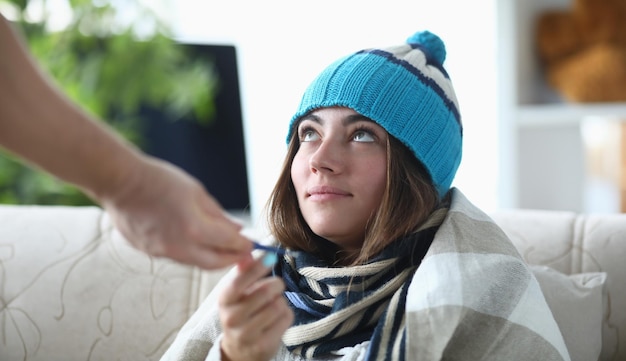 Photo une jeune femme malade avec un chapeau et un foulard est assise sous une couverture sur le canapé à la maison et reçoit des médicaments.