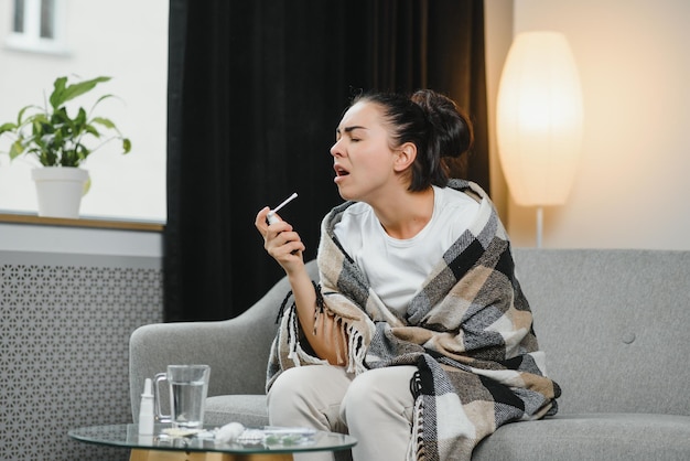 Jeune femme malade dans des vêtements chauds pulvérisant la gorge tout en étant assise sur un canapé à la maison