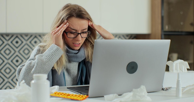 Une jeune femme malade portant des lunettes est assise au bureau, le bureau d'homa se frotte le temple, les yeux fermés tout en tombant malade et en se guérissant à la maison. Concept de soins de santé et de maladie.