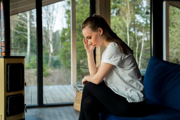 Une jeune femme malheureuse est assise à la maison sur un canapé bleu et regarde par la fenêtre en pensant à p