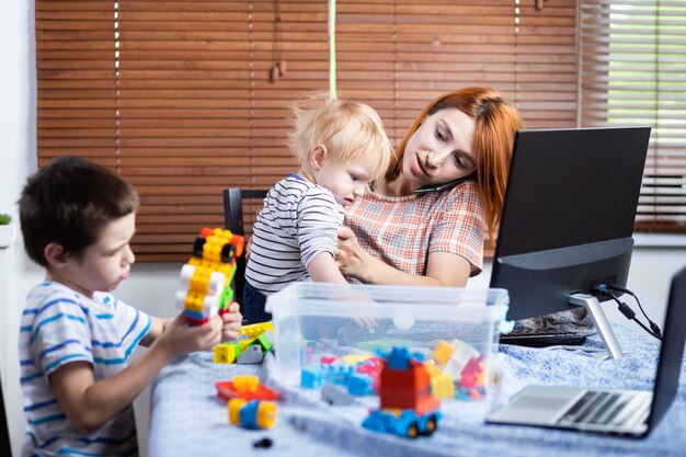 jeune femme maman parler au téléphone et essayer de travailler sur un ordinateur à distance pendant la période d'auto-isolement en lien avec la pandémie de coronavirus, soft focus.