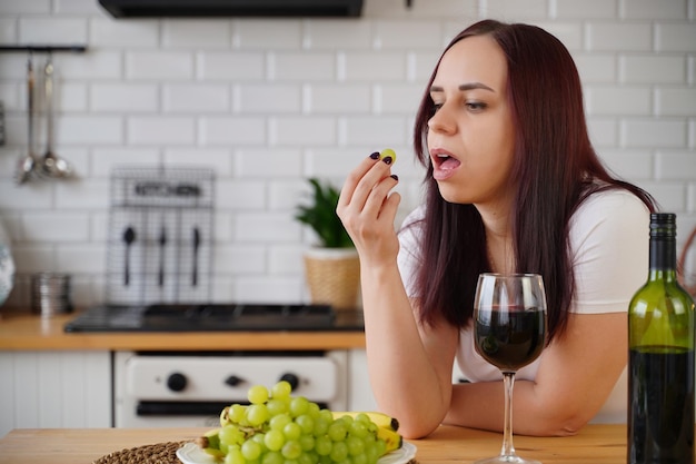 Jeune femme mange du raisin vert dans la cuisine Femelle adulte se reposant avec des fruits et de l'alcool pendant son week-end