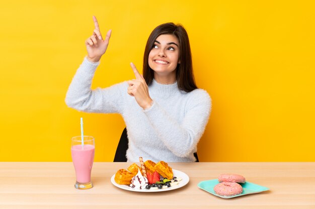 Jeune Femme Mangeant Des Gaufres Et Du Milk-shake Dans Une Table Sur Un Mur Jaune Pointant Avec L'index Une Excellente Idée