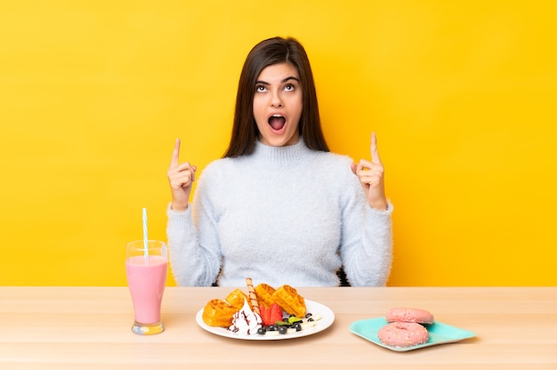 Jeune femme mangeant des gaufres et milkshake dans une table sur jaune isolé pointant avec l'index une excellente idée