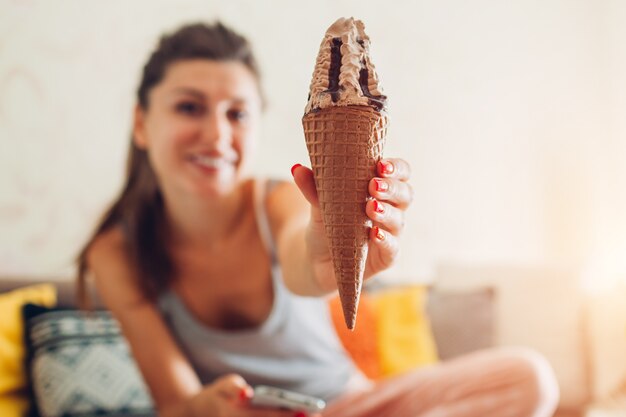 Jeune femme mangeant de la glace au chocolat en cône assis sur un canapé à la maison.