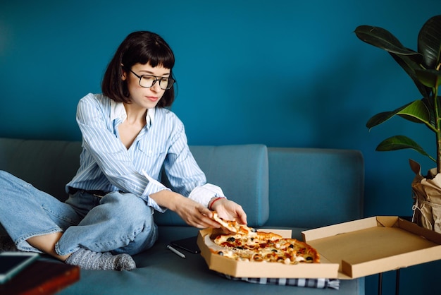Jeune femme mangeant un morceau de délicieuse pizza de la boîte, assis au canapé à la maison. Livraison de nourriture.
