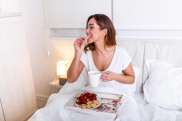 Photo jeune femme mangeant un petit déjeuner sain au lit. jeune femme prenant son petit déjeuner au lit en buvant du café. belle jeune femme souriante et mangeant des fraises.