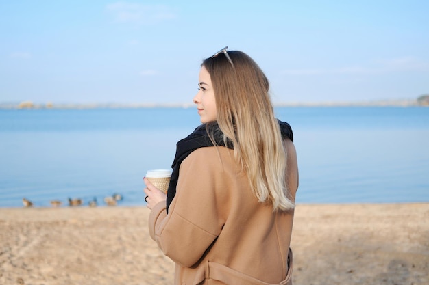 Une jeune femme en manteau marron se tient debout avec une tasse de café dans les mains et profite du temps Le concept d'harmonie et d'équilibre