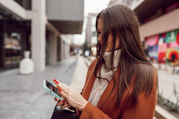 Jeune femme en manteau marron utilisant le téléphone dans les rues de la ville