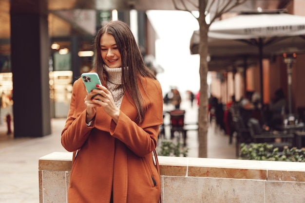 Jeune femme en manteau marron utilisant le téléphone dans les rues de la ville