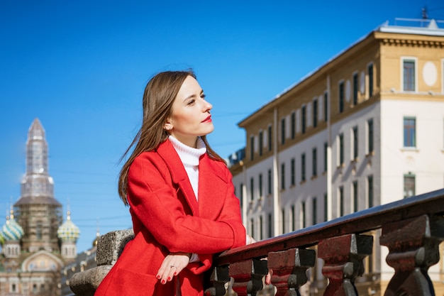 Une jeune femme en manteau rouge se promène dans la ville par une journée ensoleillée posant pour la caméra