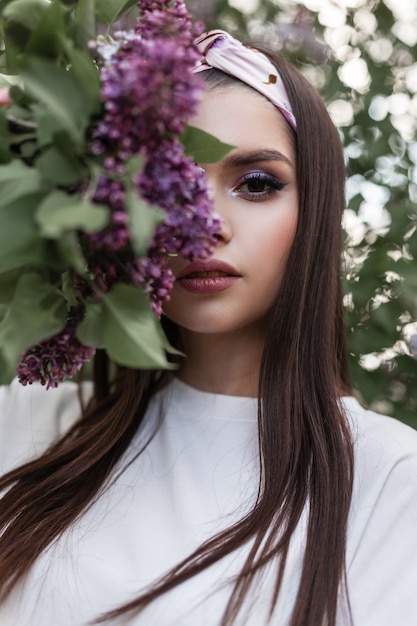 Jeune femme avec un maquillage coloré en bandana de mode dans des vêtements blancs élégants couvre le visage avec un bouquet de fleurs violettes sur fond de feuilles vertes dans le parc. Fille mignonne de portrait tendre avec des fleurs lilas.