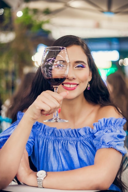 Jeune femme avec un maquillage lumineux regarde à travers un verre de vin dans un café de rue