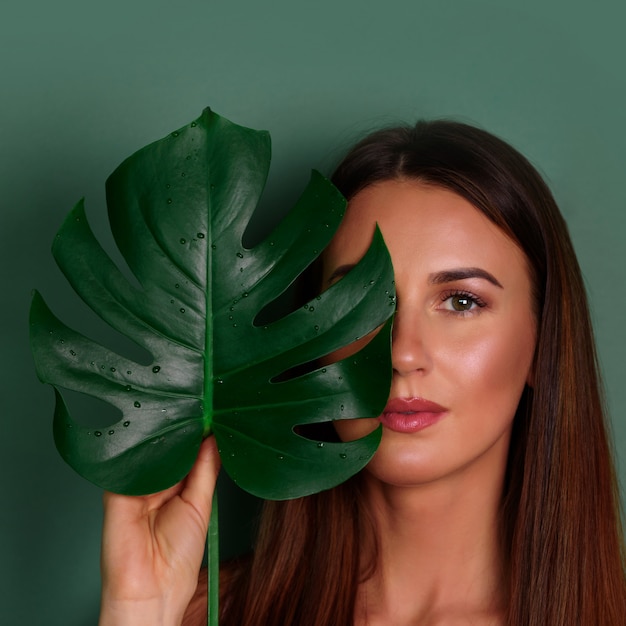 Jeune femme avec un maquillage naturel et monstera tropical feuille à la main sur fond vert avec espace de copie