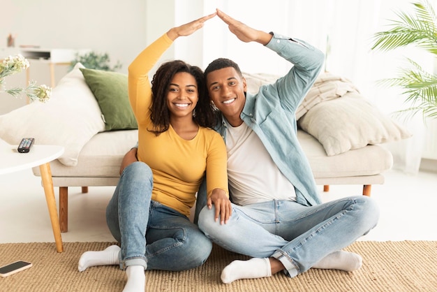 Une jeune femme et un mari afro-américains souriants s'assoient sur le sol, font le toit avec leurs mains et regardent la caméra