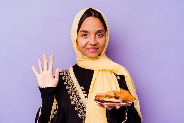 Jeune femme marocaine tenant un bonbon arabe isolé sur fond violet souriant joyeux montrant le numéro cinq avec les doigts.