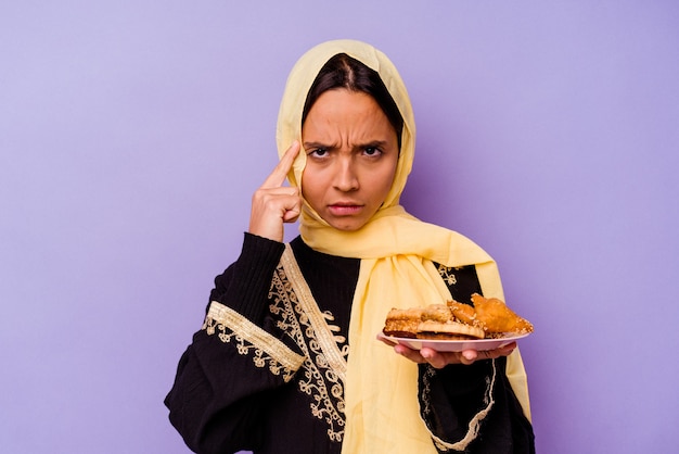 Jeune femme marocaine tenant un bonbons arabes isolé sur fond violet pointant le temple avec le doigt, pensant, concentré sur une tâche.