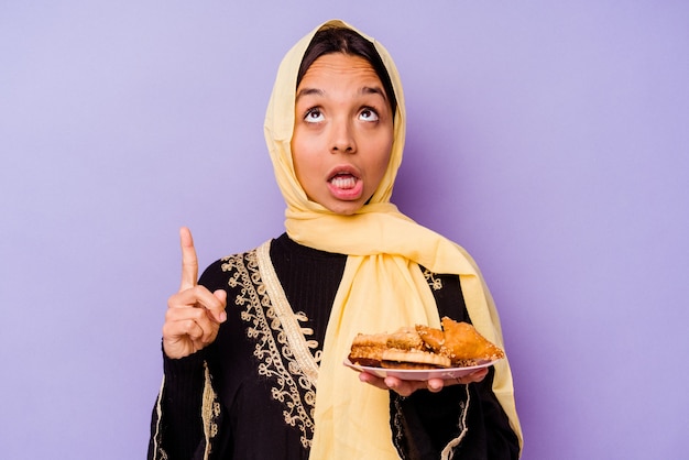 Jeune femme marocaine tenant des bonbons arabes isolés sur fond violet pointant vers le haut avec la bouche ouverte.