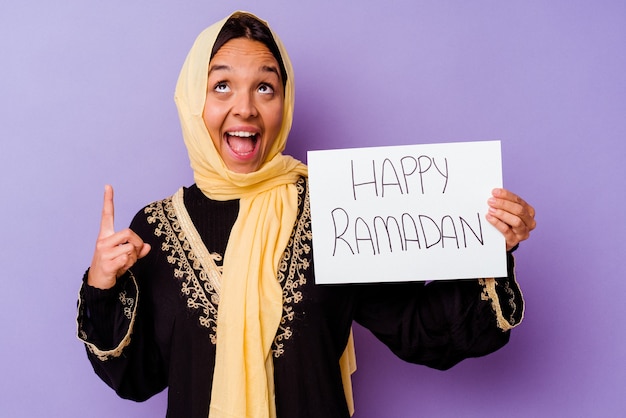 Jeune femme marocaine tenant une pancarte ramadan heureux isolé sur fond violet Jeune femme marocaine tenant une pancarte ramadan heureux isolé sur fond violet pointant vers le haut avec la bouche ouverte.