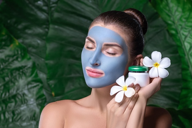 Jeune femme avec un masque d'argile bleue sur son visage. Une jeune femme tient des fleurs de frangipanier. Spa