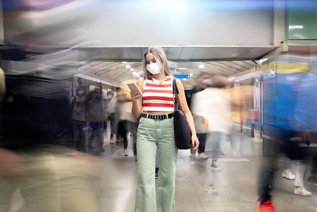 Jeune femme avec un masque blanc et un sac à main debout regardant son téléphone portable dans une station de métro