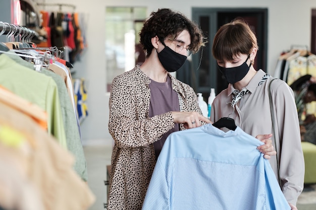 Jeune femme en masque choisissant une nouvelle chemise avec l'aide d'une vendeuse dans le centre commercial