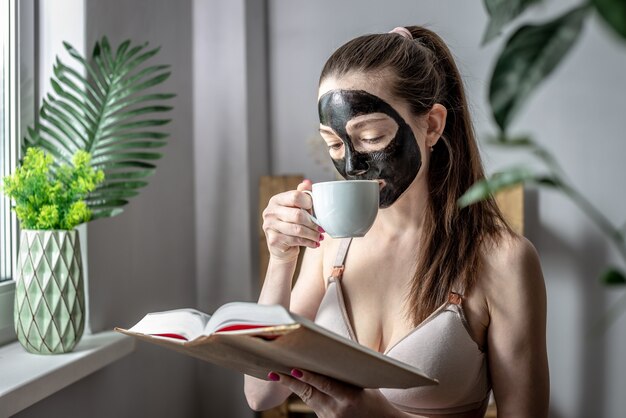 Une jeune femme avec un masque cosmétique noir sur le visage et une tasse de café lit un livre