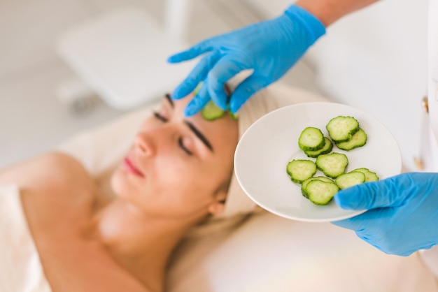 Jeune femme avec un masque facial de concombre dans un salon de beauté. Le masque cosmétique est appliqué sur le visage d'une femme. L'esthéticien tient une assiette de concombres dans ses mains