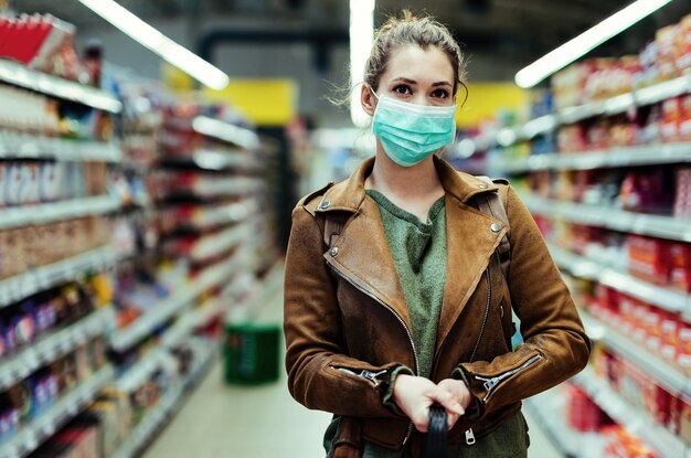 Jeune femme avec masque facial debout dans l'allée du supermarché lors de l'achat pendant la pandémie de virus